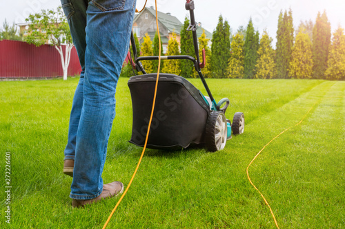 A gardener with a lawn mower is cutting green grass in the garden, in the house backyard on a sunny summer day. The legs of the worker and a lawn mower, a close-up. photo