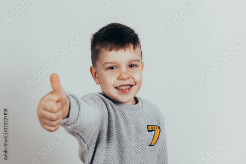 Happy child, little boy showing thumbs up gesture in gray sweater against gray background. Space for your text.