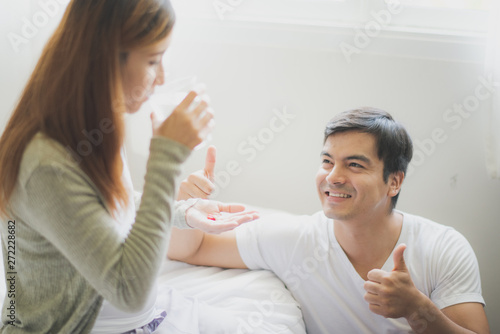 Young pregnant Asian woman and her husband are sitting on the white bed which is smiling felling happy at home. Man is giving drug to woman. To be new Mother and Father, pregnant woman concept.