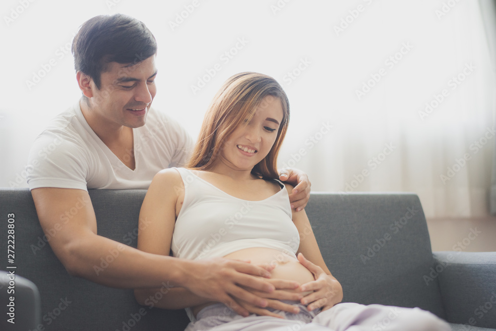 Young pregnant Asian woman is sitting on sofa with her husband are looking at her belly near window which felling happy at family's home. To be new Mother and Father, pregnant woman concept. Young pregnant Asian woman is sitting on sofa with her husband are looking at her belly near window which felling happy at family's home. To be new Mother and Father, pregnant woman concept.