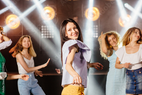 Pretty brunette female clubber dancing surrounded by her friends and looking at camera with smile. Bright lighting with spotlights.