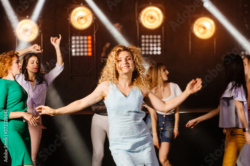 Curly blond female clubber dancing surrounded by her friends and looking at camera with smile during holidays party relaxing celebrating dancing at party in night club together.