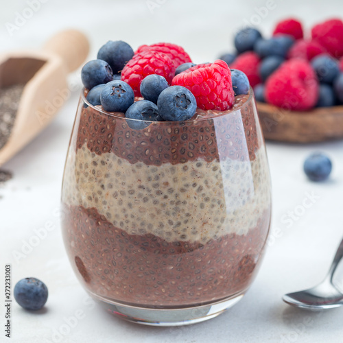 Layered chocolate and peanut butter chia seed pudding in a glass, garnished with raspberry and blueberry, square format