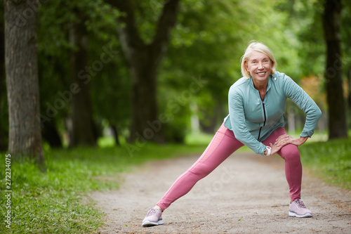 Mature cheerful blonde female in activewear stretching legs while doing physical exercises on road in city park among green trees © Seventyfour