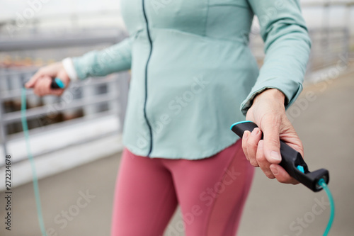 Hands of mature sportswoman holding skipping rope while jumping during workout in urban environment
