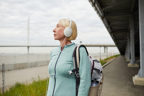 Mature blonde active female traveler in sportswear listening to music in headphones while chilling out in urban environment