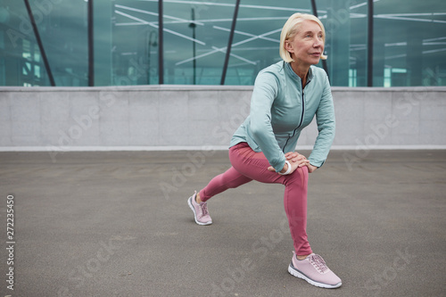 Pretty active mature woman in sportswear doing exercisie for leg muscles on concrete by contemporary building outdoors