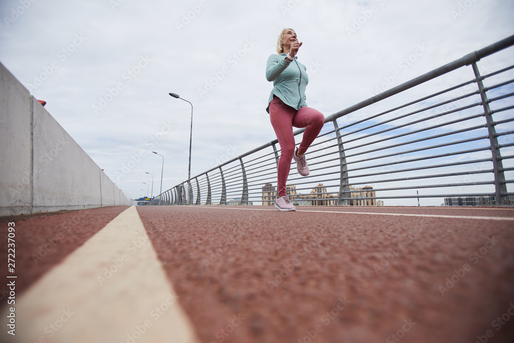 Contemporary active mature female in sportswear jogging along bridge border while training on stadium outdoors