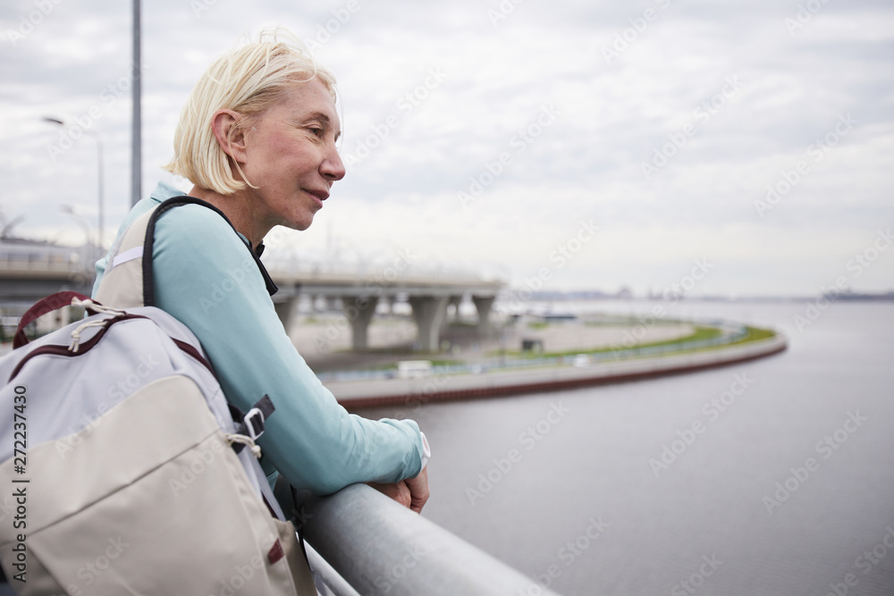 Contemporary mature blonde female backpacker in activewear standing by riverside while enjoying solitude outdoors