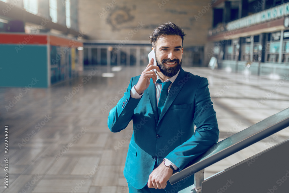 Young gorgeous smiling Caucasian businessman in blue suit leaning on railing and using smart phone at train station.