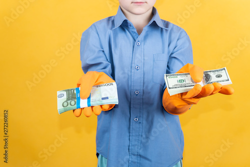 A child in a blue shirt and washing gloves is holding a pack of dollars in one hand and a pack of euro notes in the other. The concept of financial independence with clean hands.