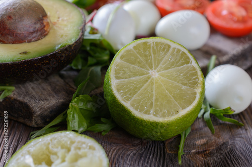 Half of green lime and other ingredients for salad as ripe and healthy avocado with a bone, greens, quail eggs and tomato cherries on a wooden table, closeup