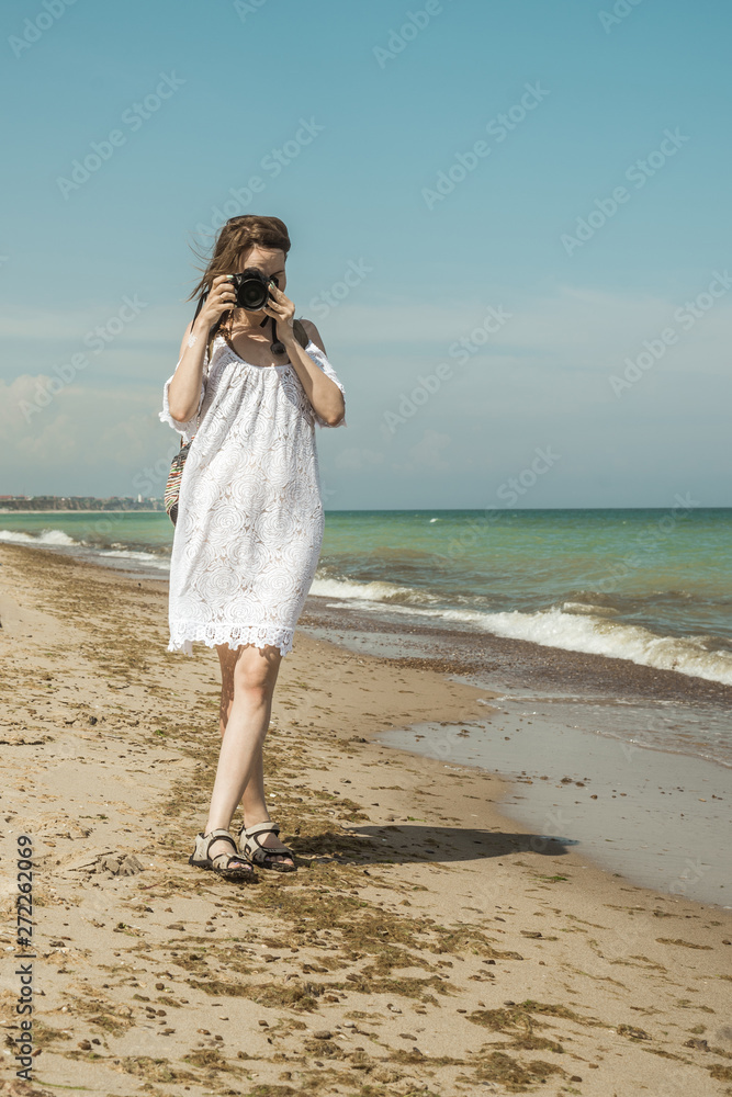 Woman photographer at the sea. A middle-aged woman in a white dress with a camera goes along the coast. Summer tourism, photographing attractions on vacation. Woman photographer at the sea. A middle-aged woman in a white dress with a camera goes along the coast. Summer tourism, photographing attractions on vacation.