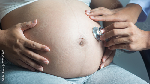 close up of gynecologist doctor with stethoscope listening to pregnant woman baby heartbeat at hospital