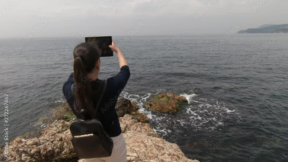 A tourist with a backpack behind shoots a video of sea waves on his tablet A tourist with a backpack behind shoots a video of sea waves on his tablet