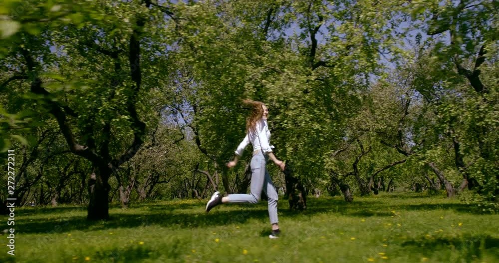 A young beautiful slender girl in jeans and a white shirt runs in the field past the trees and bushes on a summer day. Around it green the grasses and green trees. A young beautiful slender girl in jeans and a white shirt runs in the field past the trees and bushes on a summer day. Around it green the grasses and green trees.