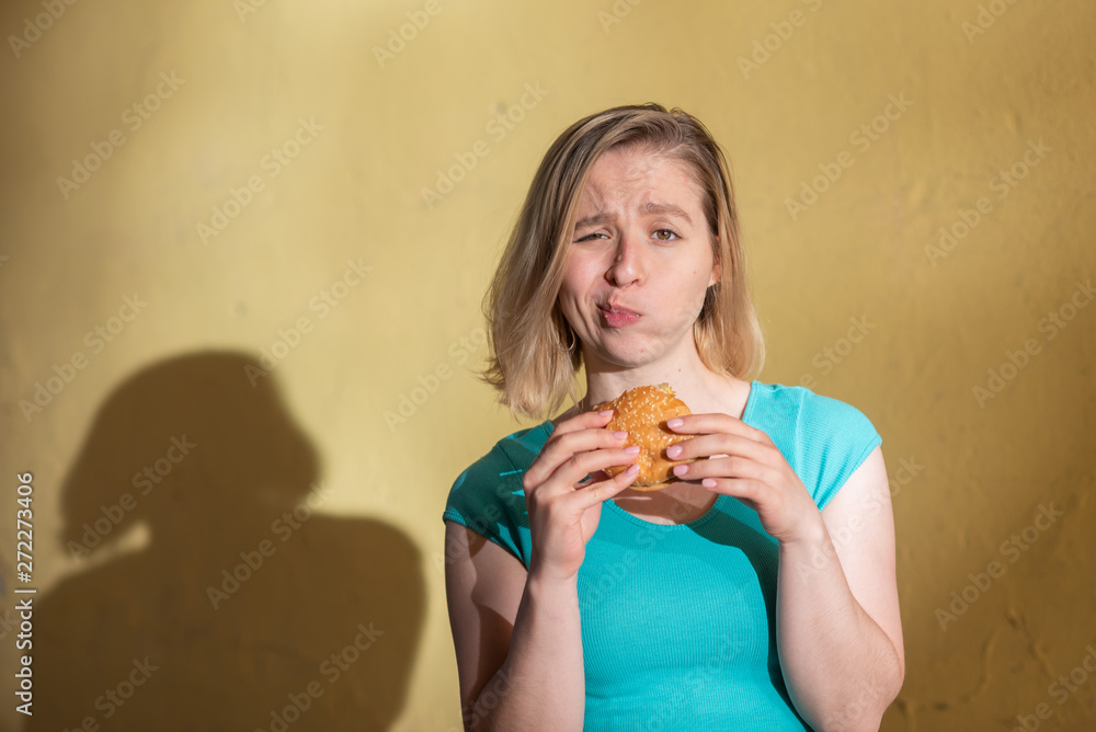 Portrait of a cute girl in a green dress walking outdoors and eating a burger on a warm summer day. Beautiful blonde is enjoying fast food at the yellow wall outside. Wrong food habits.