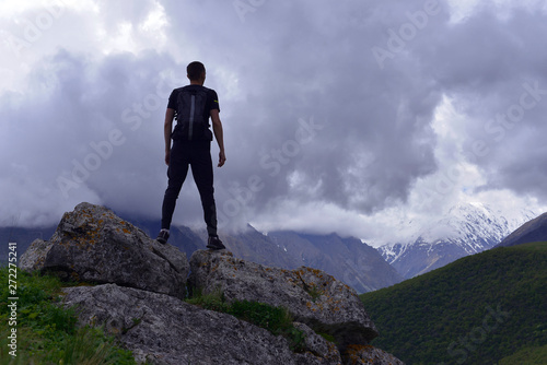 A young man with a backpack stands high on a hill on a large stone and looks at the dark blue clouds before the rain.