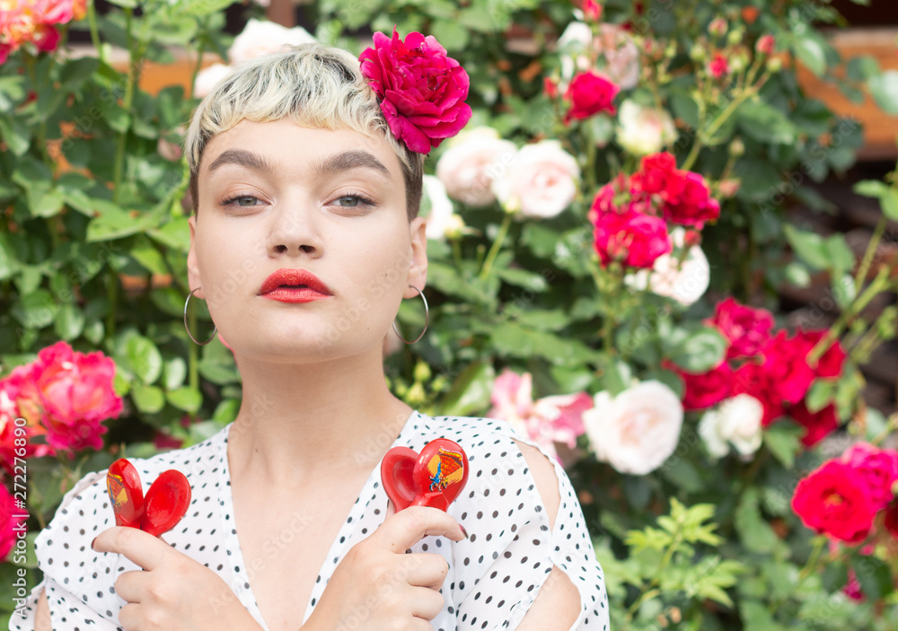 Naklejka premium A beautiful young blonde girl in a white blouse and with a rose in her hair expresses emotions in a rose garden on the background of an amazing summer. The girl plays with red castanets.