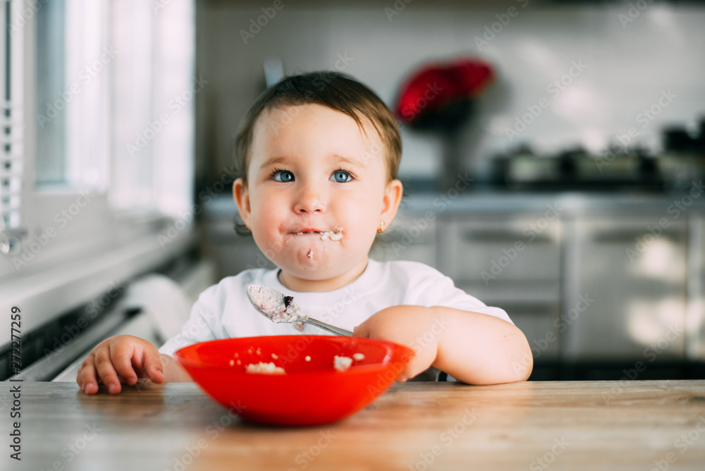 Funny little girl in the kitchen during the day eating oatmeal porridge from a red plate all dirty Funny little girl in the kitchen during the day eating oatmeal porridge from a red plate all dirty