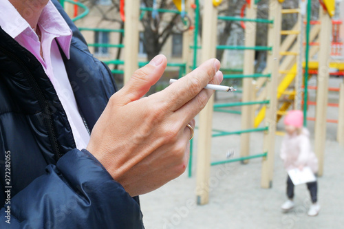 man smokes near children playground, hand with a cigarette and smoke closeup, bad habits concept