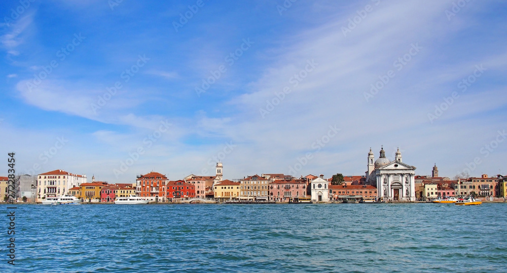 a long seafront panoramic cityscape view of the salute area of venice with historic buildings along the waterfront and a bright blue sunlit sky