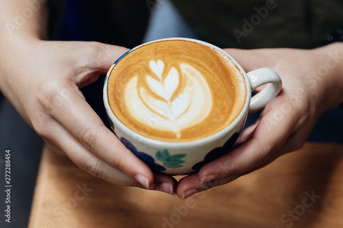 Top view of hands holding a Latte art heart shape served in ceramic cup on wooden table.