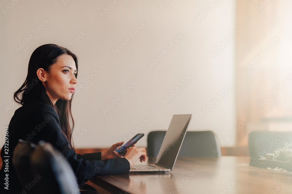 Young attractive emotional girl in business-style clothes sitting at a desk on a laptop and phone in the office or auditorium Young attractive emotional girl in business-style clothes sitting at a desk on a laptop and phone in the office or auditorium