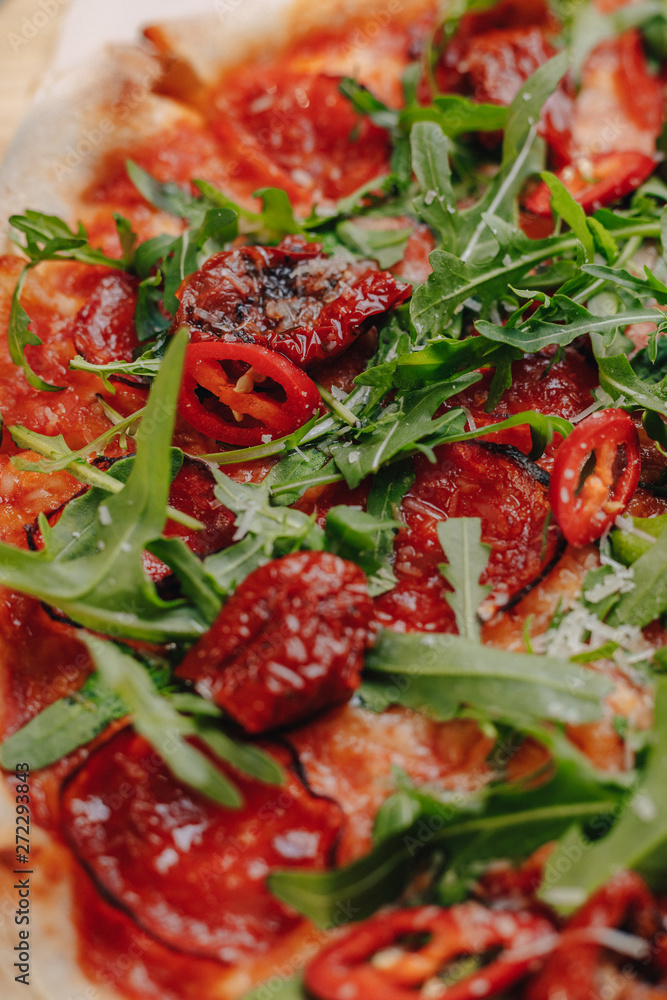Neapolitan spicy pizza with ham, cheese, arugula, basil, tomatoes, pepperoni pepper sprayed with cheese on a wooden board on a tablecloth in a cell with a place for text Neapolitan spicy pizza with ham, cheese, arugula, basil, tomatoes, pepperoni pepper sprayed with cheese on a wooden board on a tablecloth in a cell with a place for text