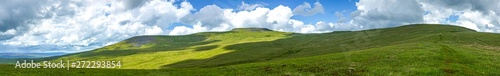 A panoramic view of a mountain with grassy green slope under a majestic blue sky and white clouds