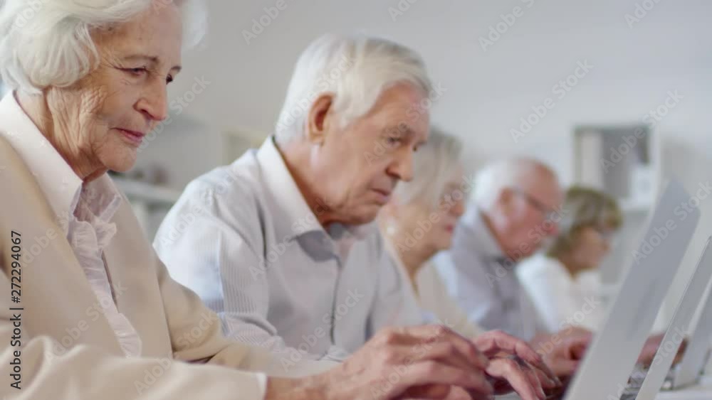 Tilt up shot with side view of focused elderly men and women with grey hair sitting at desks and typing on laptops during computer class