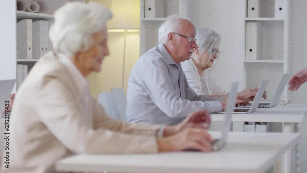 Tracking shot of male technology teacher with beard walking among desks of elderly people using laptops and monitoring their progress Tracking shot of male technology teacher with beard walking among desks of elderly people using laptops and monitoring their progress