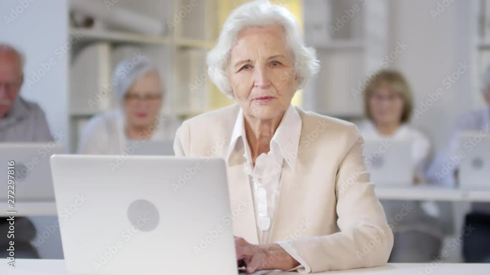 Portrait shot of serious elderly woman with grey hair sitting at desk and using laptop in computer class, then looking at camera Portrait shot of serious elderly woman with grey hair sitting at desk and using laptop in computer class, then looking at camera