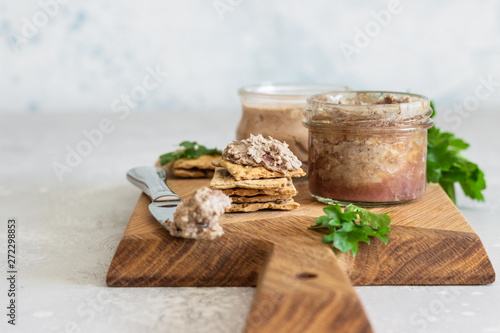 Fresh homemade liver pate in a glass jar with multigrain crackers and parsley on a wooden background.