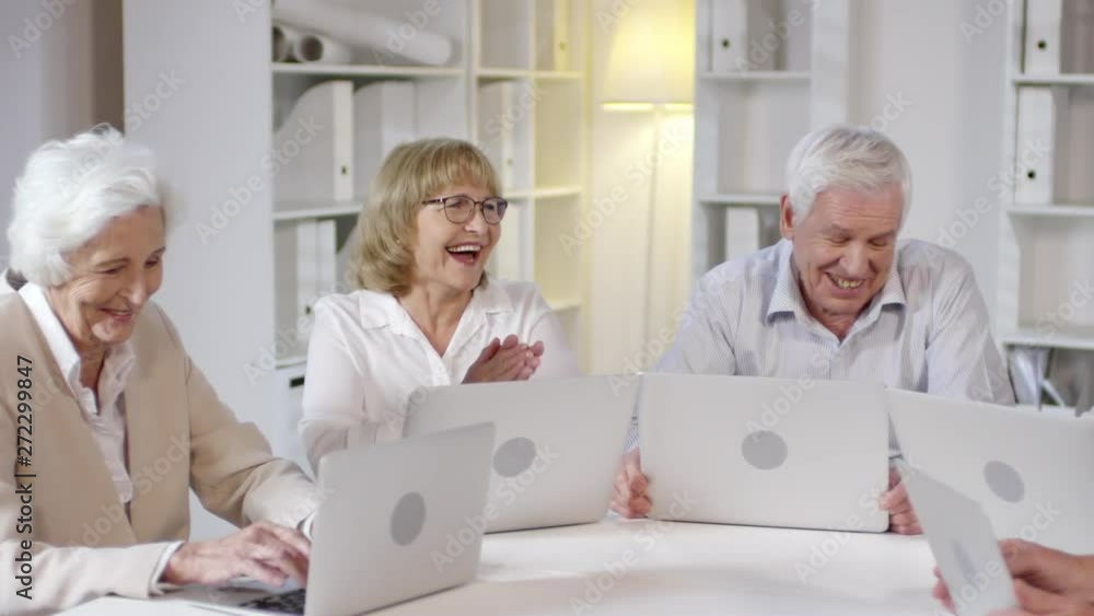 PAN shot of happy elderly men and women with laptops sitting at desk and chatting about something PAN shot of happy elderly men and women with laptops sitting at desk and chatting about something