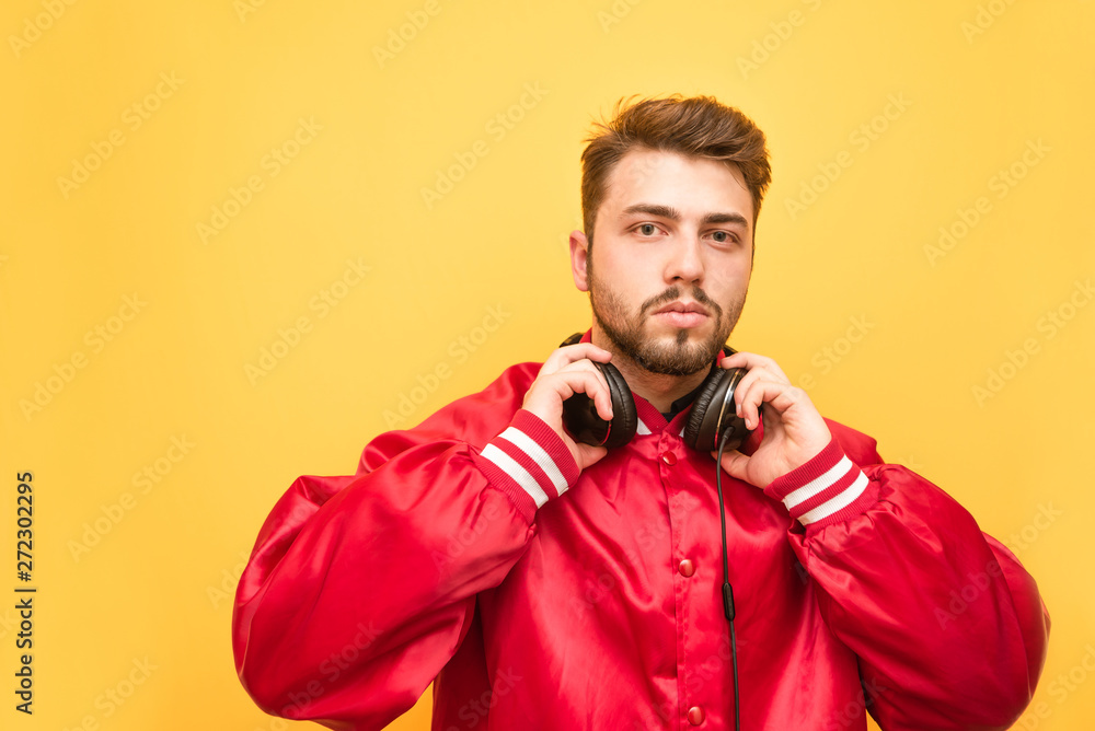 Portrait of a bearded man in his headphones and a red jacket on a yellow background, looking up at the camera. Adult student with headphones on the neck is isolated. Portrait of a bearded man in his headphones and a red jacket on a yellow background, looking up at the camera. Adult student with headphones on the neck is isolated.