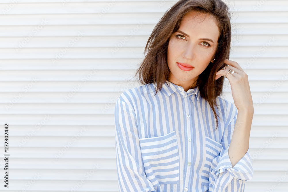 Close portrait of young stylish beautiful brunette woman, wearing blue striped shirt, posing outdoors on a background of white wall. Street fashion. Space for text. Close portrait of young stylish beautiful brunette woman, wearing blue striped shirt, posing outdoors on a background of white wall. Street fashion. Space for text.