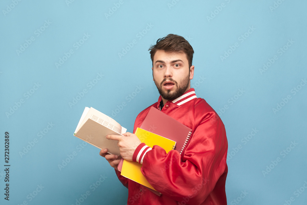 Amazed young man with a beard and books in his hands, wearing a red jacket, looks into the camera with shocking face. Portrait of astonished student husband. Studying at the University of Concepts. Amazed young man with a beard and books in his hands, wearing a red jacket, looks into the camera with shocking face. Portrait of astonished student husband. Studying at the University of Concepts.