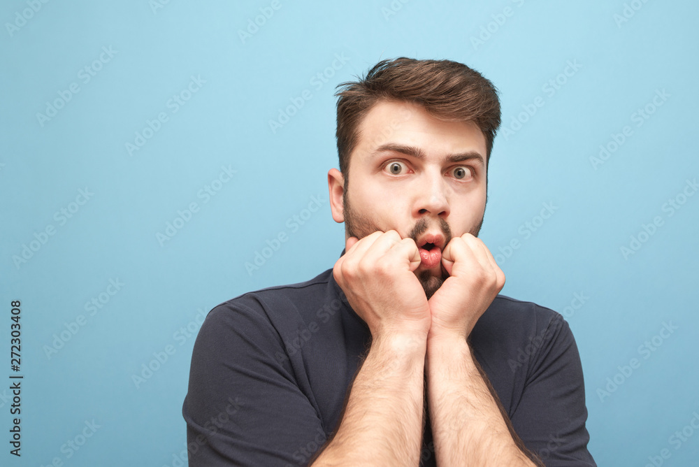 Close-up portrait of a frightened man with a beard on a blue background looks shocked at the camera. Wear a dark shirt. Isolated. Close-up portrait of a frightened man with a beard on a blue background looks shocked at the camera. Wear a dark shirt. Isolated.