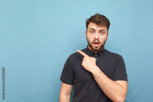 Surprised adult man with a beard and a dark shirt shows a finger toward the blank face and shocked looks from the side. Isolated. Copy space