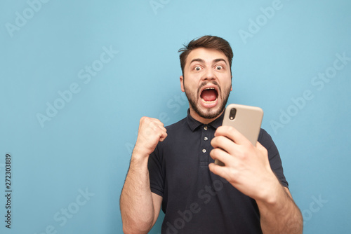 Emotional bearded man holds the smartphone in his hands, looks at the camera and rejoices. Man with a smartphone in his hands rejoices in success, isolated on a blue background. Copyspace