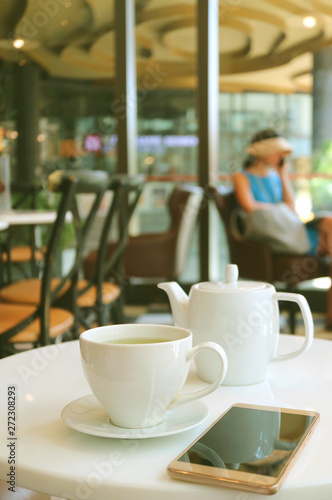 Cup of Hot Green Tea and White Teapot with a Mobile Phone on Cafe's Table 