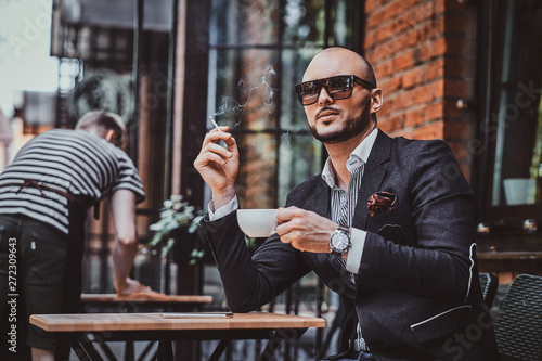 Pensive modern man is smoking a cigarette outside of coffeshop while drinking his cup of coffee. photo