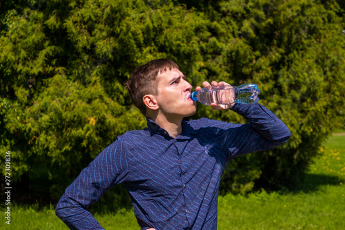 portrait of men drinking water from a bottle, in blue shirt standing outside in park