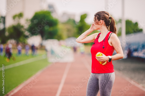 The topic is sport and health. A young Caucasian woman in training in sportswear is talking using a mobile phone, the hand is ear phone, at the sports stadium, the weather is sunny. © Elizaveta