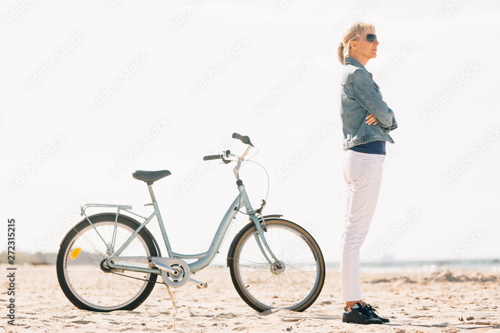 Fototapeta premium Pretty blonde girl in white pants and denim coat standing on the beach with bicycle. Atrractive woman relaxing near the sea after bike ride
