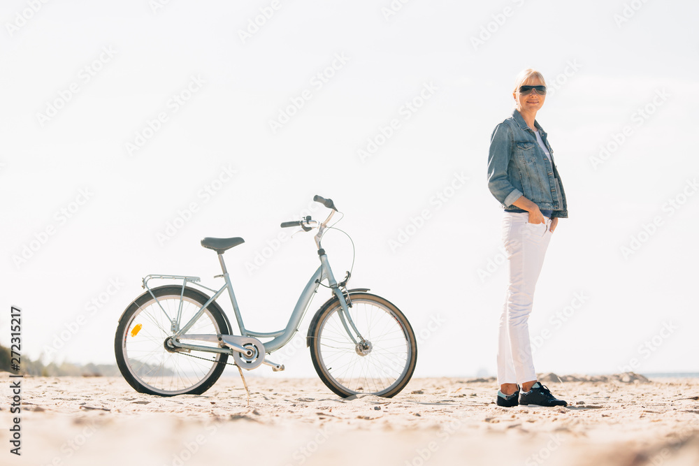 Fototapeta premium Pretty blonde girl in white pants and denim coat standing on the beach with bicycle. Atrractive woman relaxing near the sea after bike ride