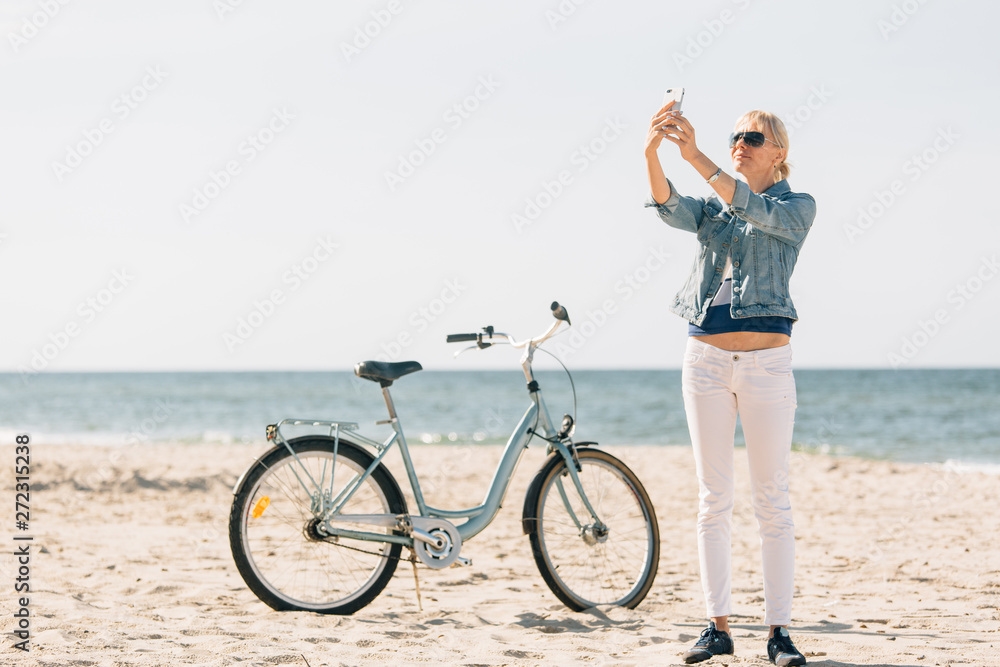Naklejka premium Pretty blonde girl in white pants and denim coat taking selfie on the beach with bicycle. Atrractive woman relaxing near the sea after bike ride