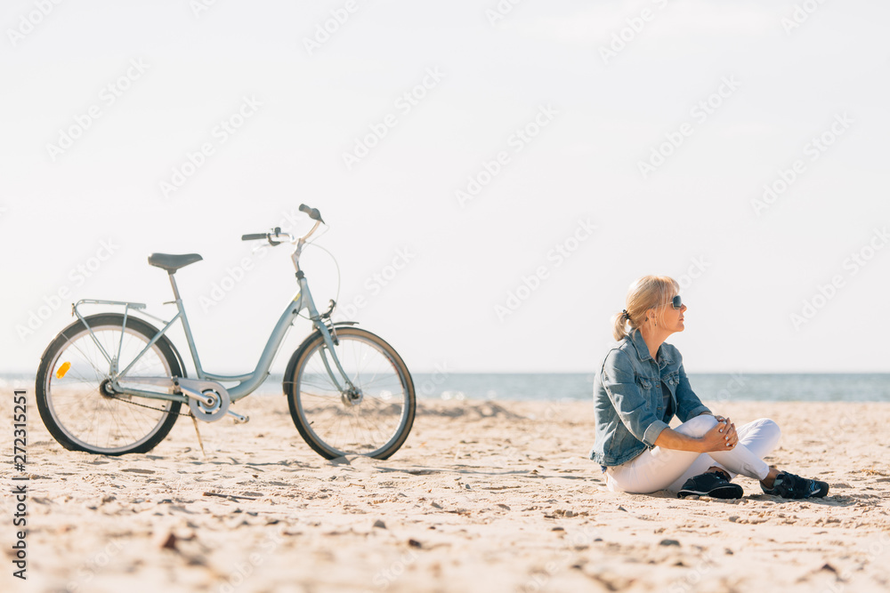 Pretty blonde girl in white pants and denim coat sitting on the beach near bicycle. Atrractive woman relaxing near the sea after bike ride