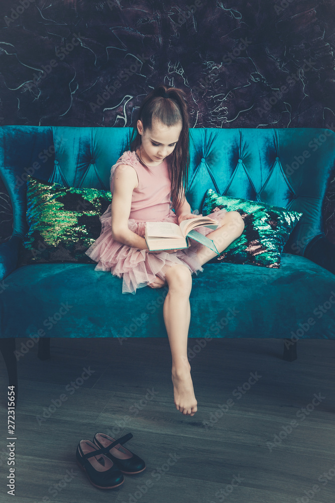 Cute girl with brown hair in a pink dress sitting on a blue sofa and reading a book, eyes downcast.	